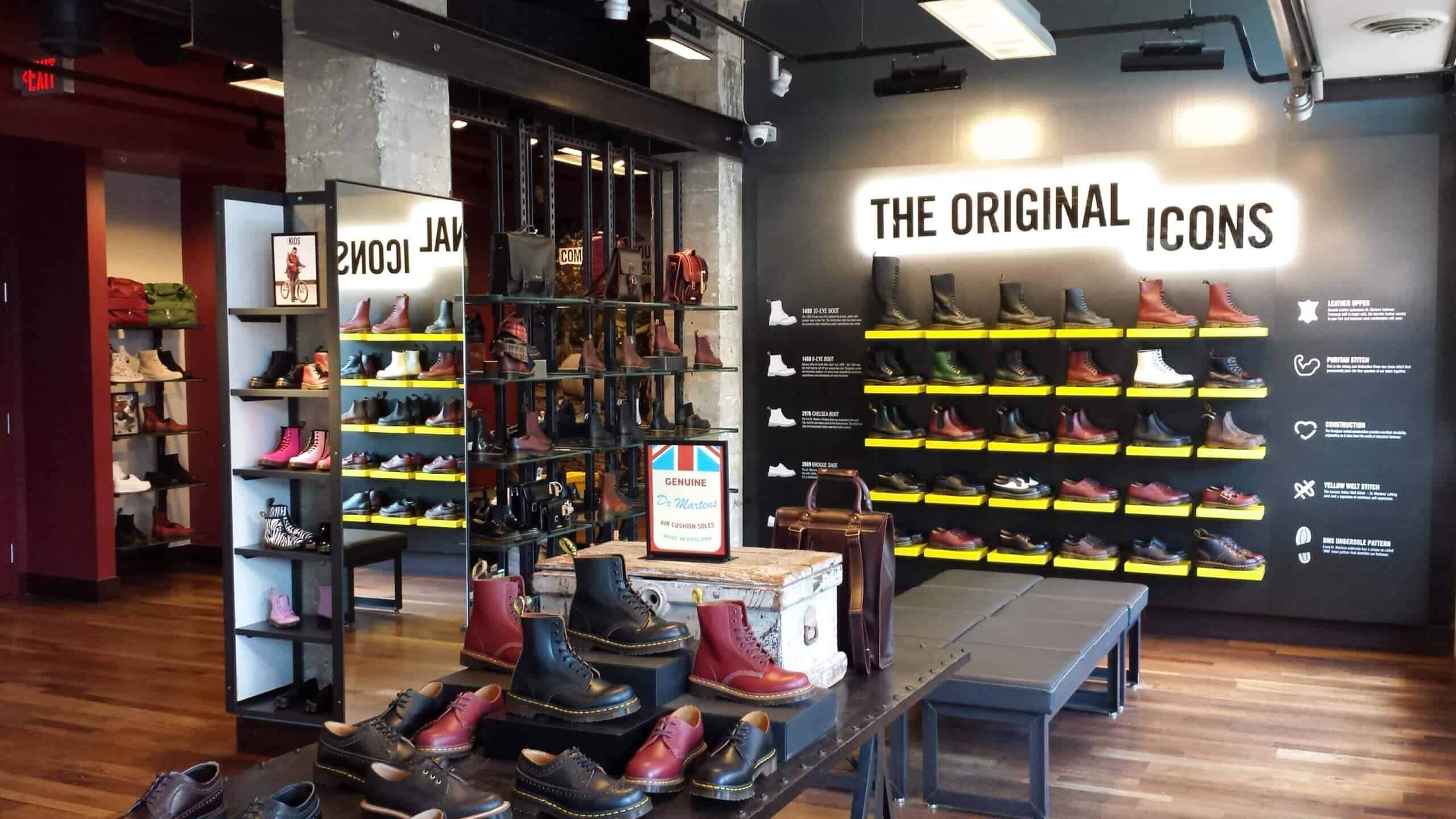 Interior of a Dr. Martens retail store showcasing various iconic boot models on black and yellow wall shelves, with additional footwear and leather goods displayed on tables and side shelves.