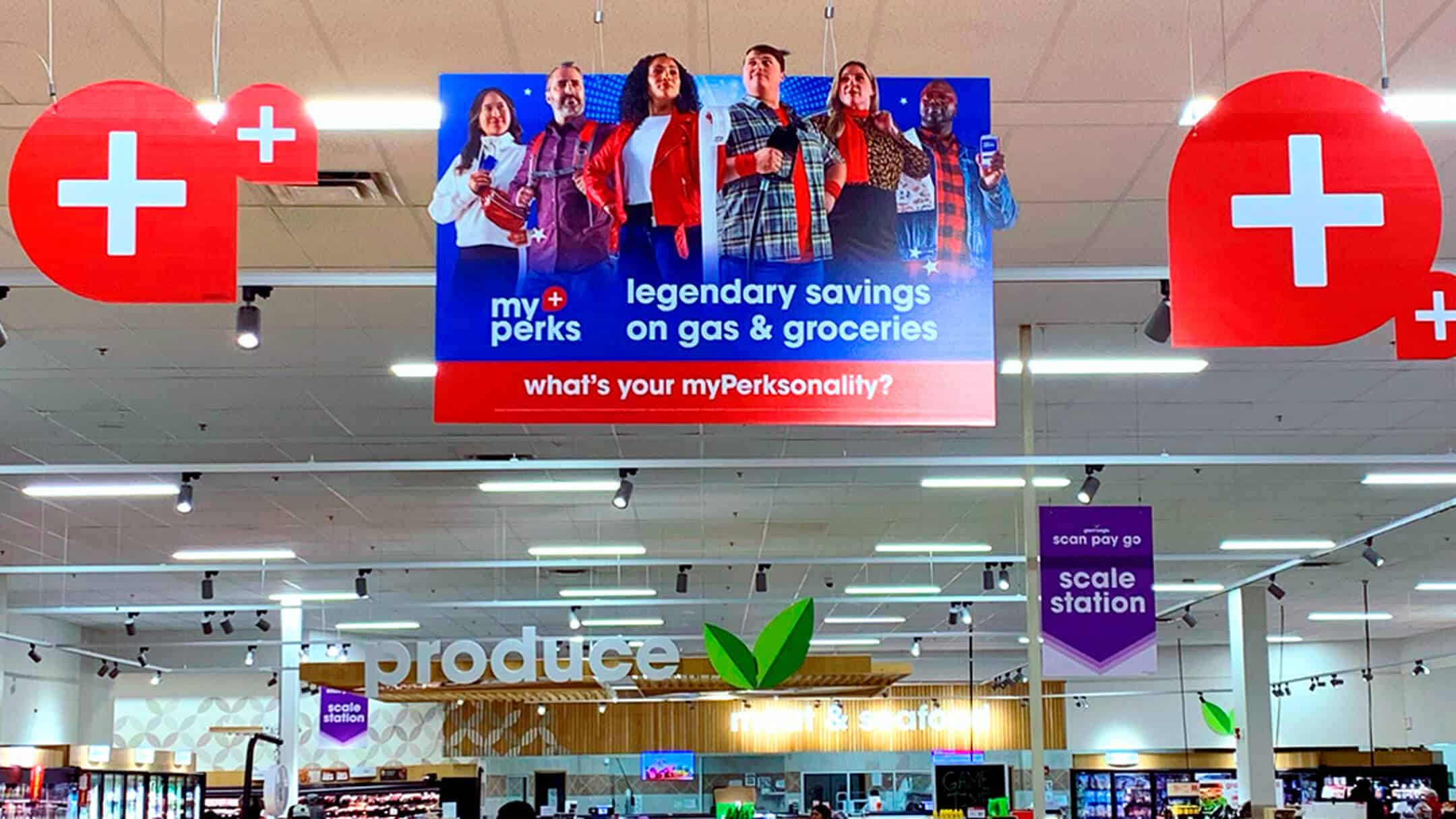 Large hanging sign in grocery store advertising myPerks savings with six diverse people and red plus icons on either side.
