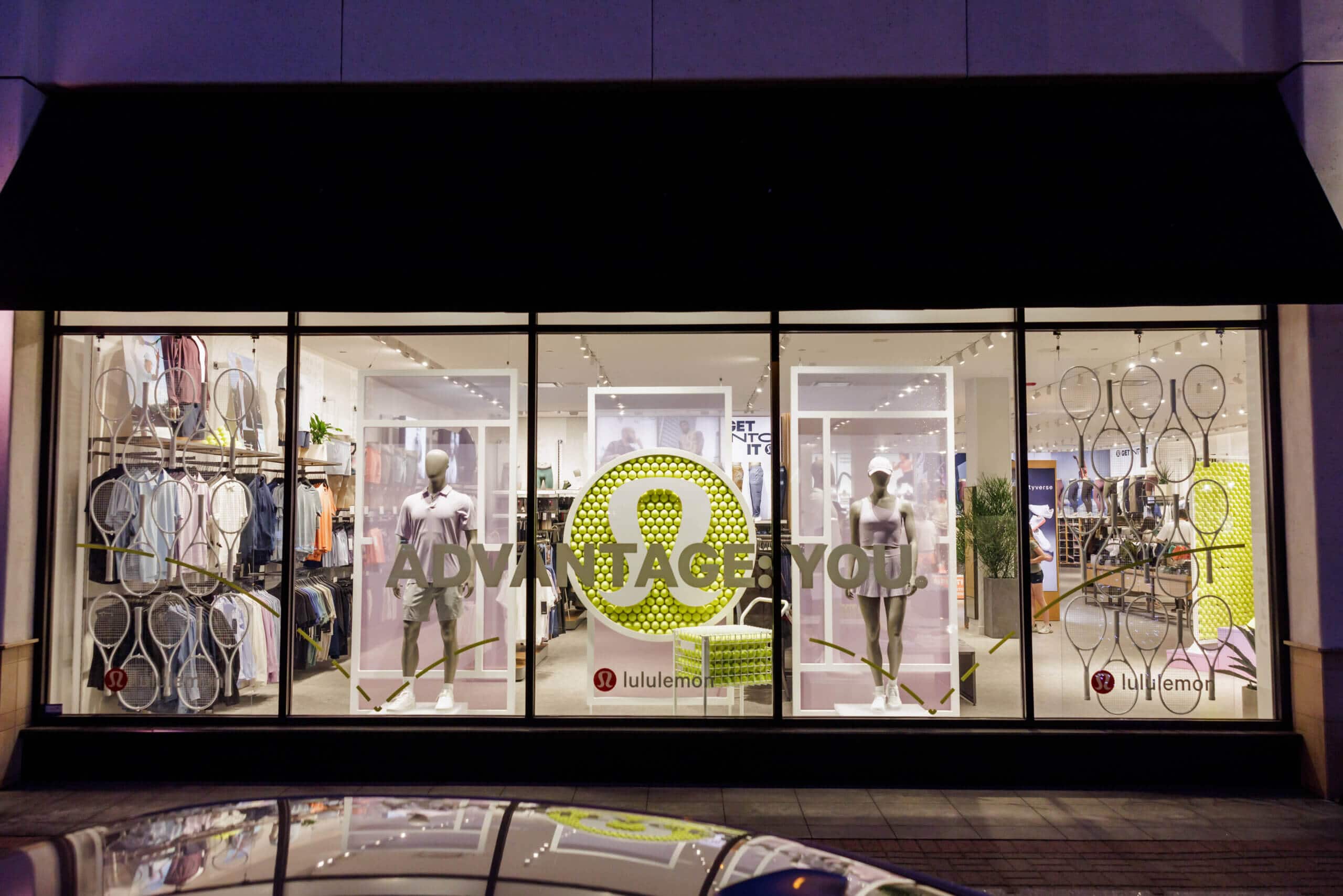 Lululemon storefront window display with tennis theme, featuring mannequins, tennis rackets, and a wall of tennis balls forming the Lululemon logo.