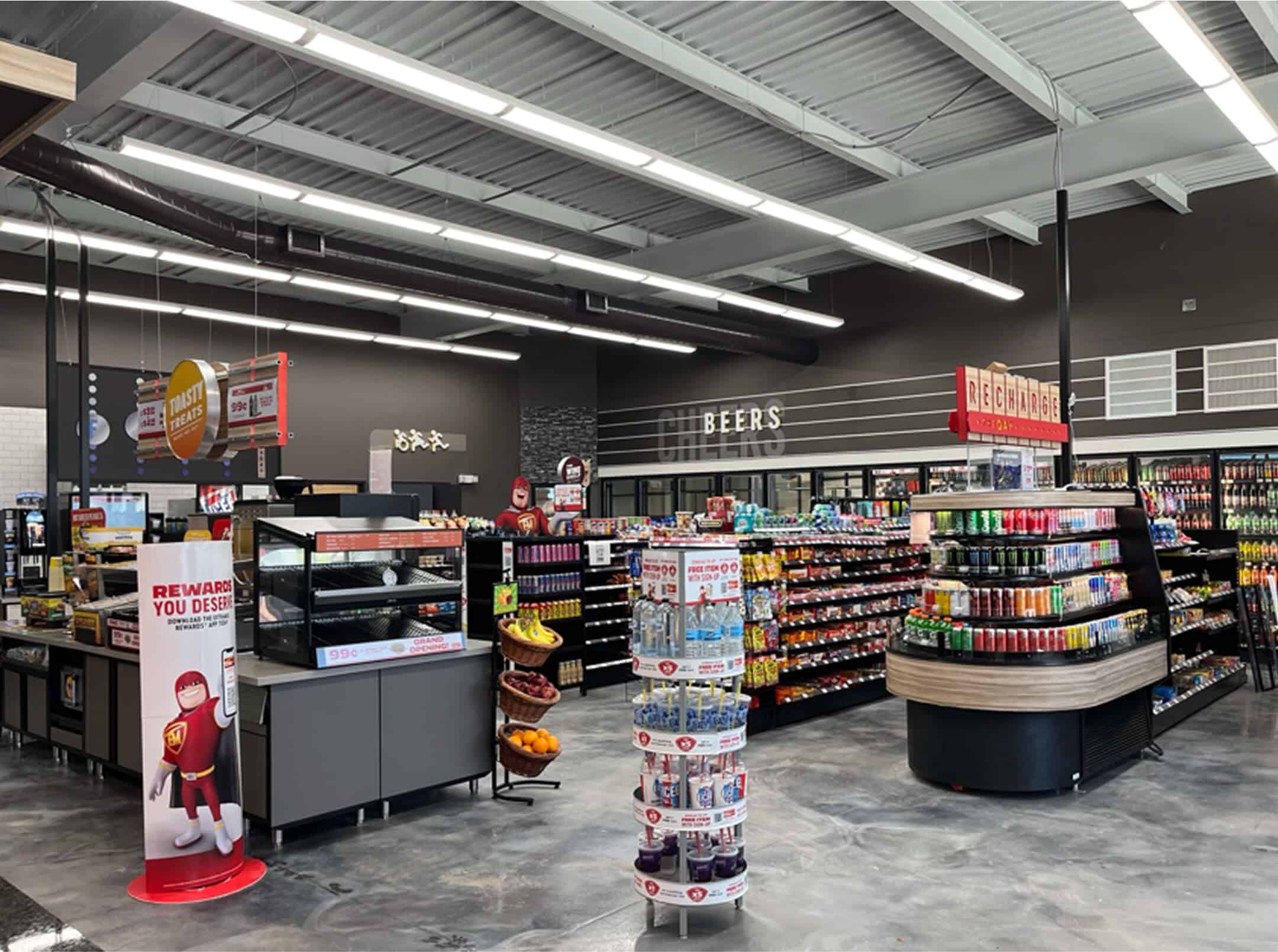 Interior of ExtraMile convenience store with beverage coolers, snack shelves, promotional displays, and a “Recharge the Day” drink station.