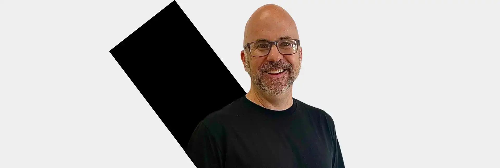 Smiling Kevin Peters wearing a black t-shirt, standing in front of a white background with a tilted black square behind him.
