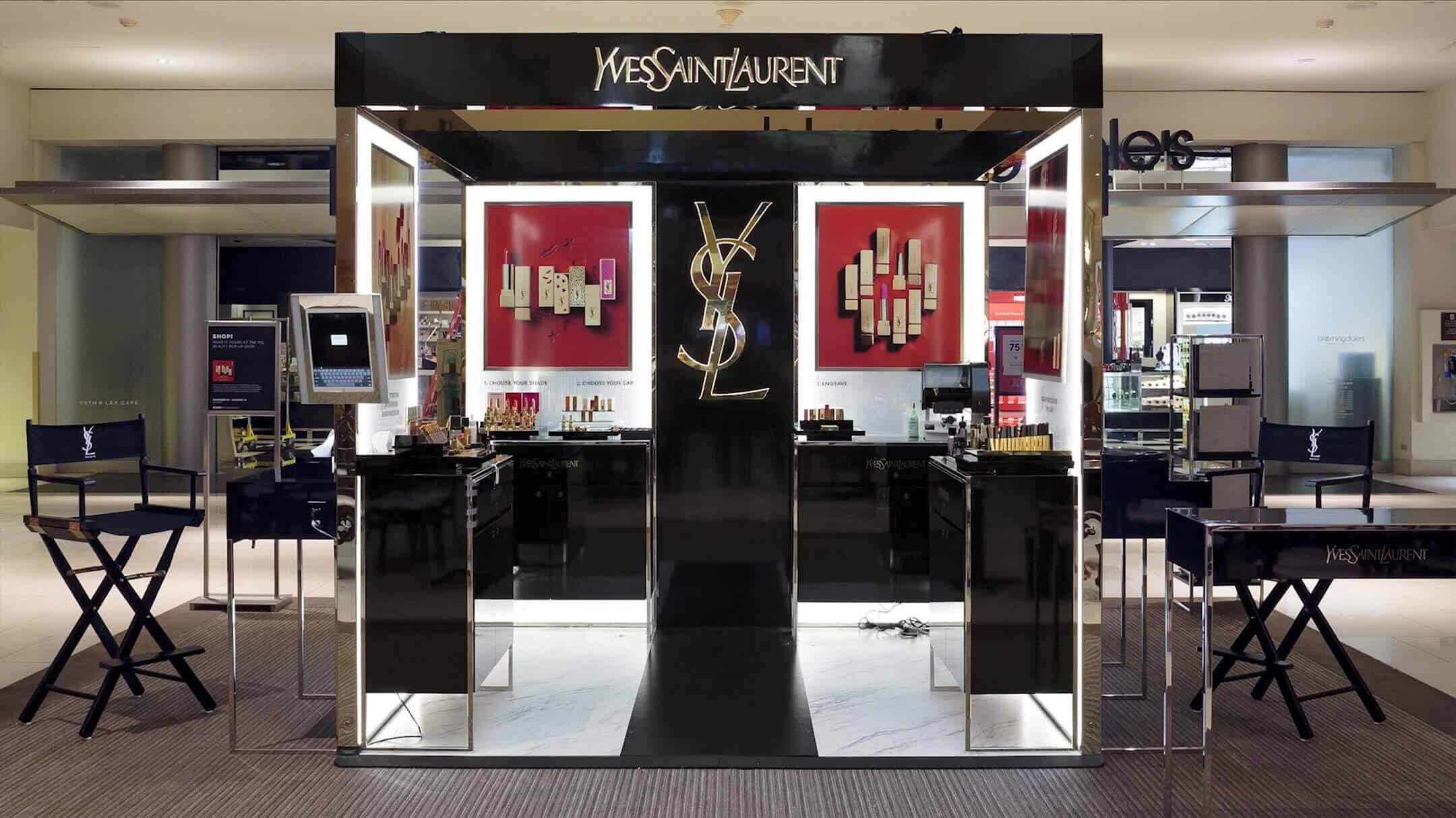Yves Saint Laurent (YSL) cosmetic kiosk inside a department store featuring sleek black counters, makeup displays, and YSL-branded chairs.