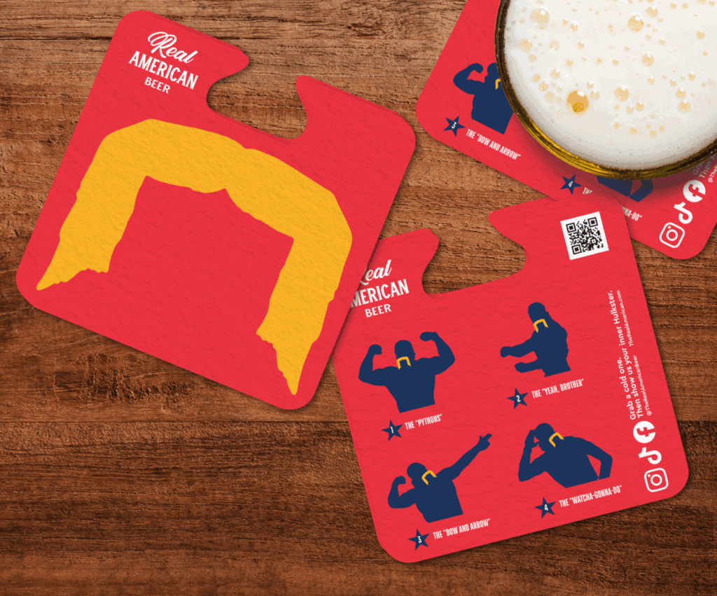 Close-up of red Real American Beer coasters with yellow mustache and flexing arm graphics on a wooden table.